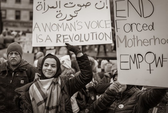 Picture shows a woman in a hijab in the middle of a protest smiling as she holds a cardboard sign which reads 'A woman's voice is a revolution'